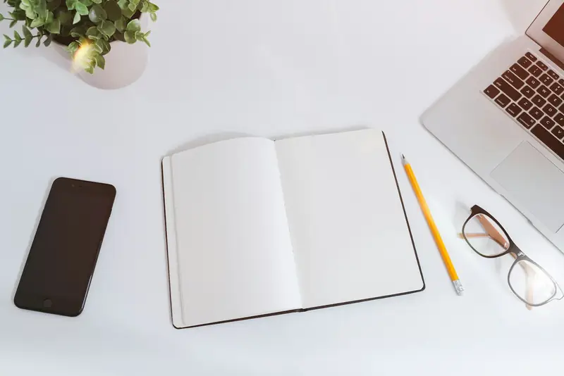 Open notebook, phone, laptop and glasses on a white desk